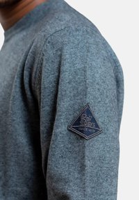 Maglione grigio a maglia con scollo rotondo e una toppa triangolare blu sul maniche sinistra con testo bianco. Texture morbida, design aderente.