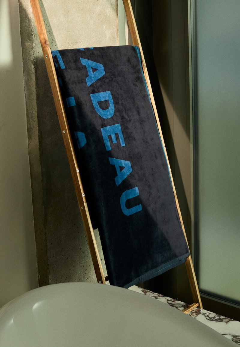 Mörkblå handduk med strukturerad yta, med stora turkosa bokstäver, hängande på en trästege mot en minimalistisk bakgrund.