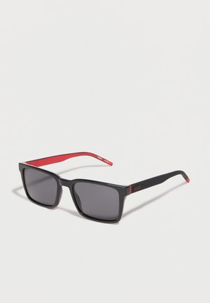 Μαύρα ορθογώνια γυαλιά ηλίου με σκουρόχρωμους φακούς, υφασμένα μαύρα πλαίσια και εσωτερικά κόκκινα πλαίσια με την επωνυμία "HUGO", εμφανίζονται σε ανοιχτό φόντο.