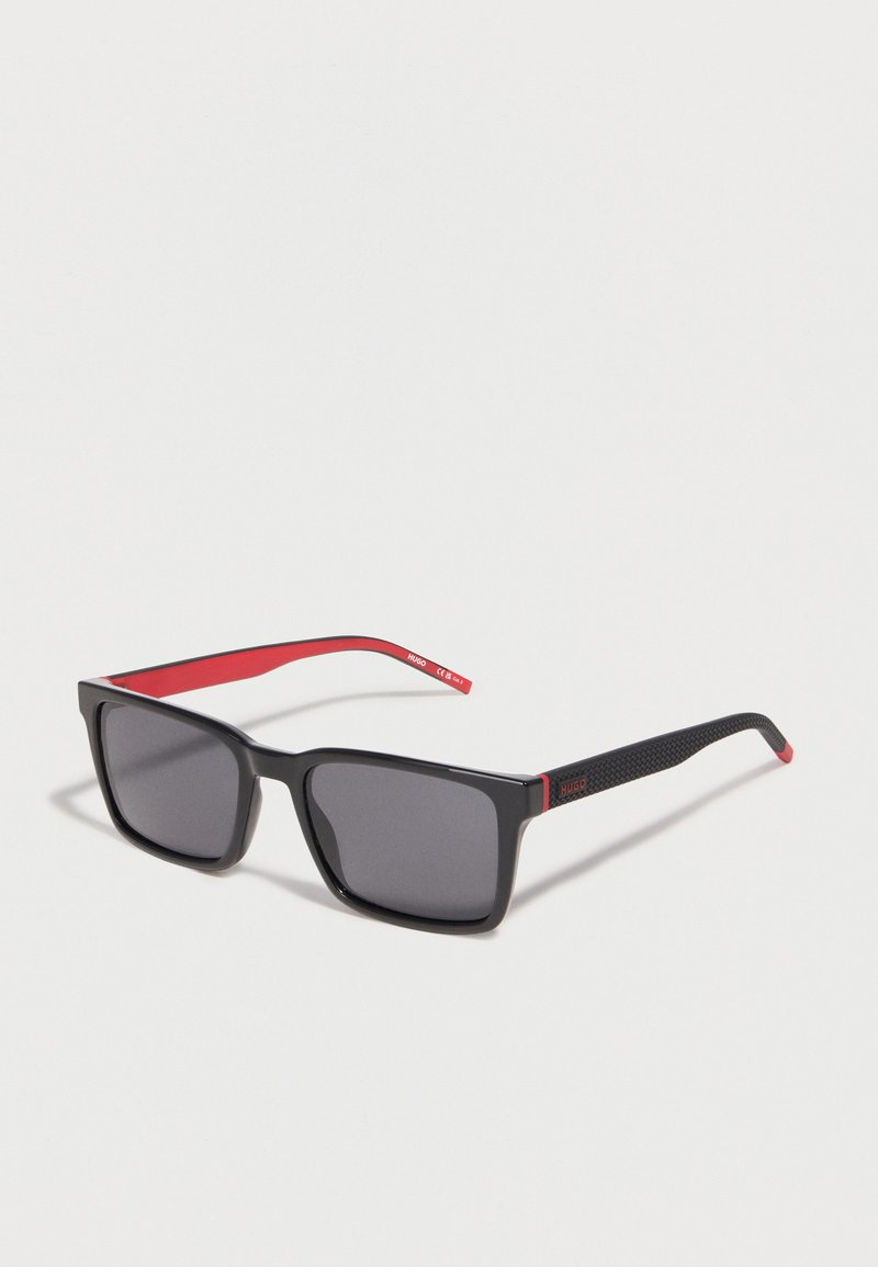Μαύρα ορθογώνια γυαλιά ηλίου με σκουρόχρωμους φακούς, υφασμένα μαύρα πλαίσια και εσωτερικά κόκκινα πλαίσια με την επωνυμία "HUGO", εμφανίζονται σε ανοιχτό φόντο.