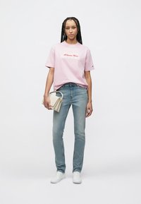 Hellrosa Baumwoll-T-Shirt mit grafischem Text, kombiniert mit hellblauen Jeans und weißen Turnschuhen. Das Modell trägt eine beige Tasche.