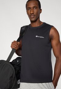 Μαύρο αθλητικό τοπ χωρίς μανίκια με λευκό λογότυπο "Champion". Το μοντέλο κρατά μια μαύρη τσάντα με γκρι λεπτομέρειες. Λείο ύφασμα και άνετη εφαρμογή.
