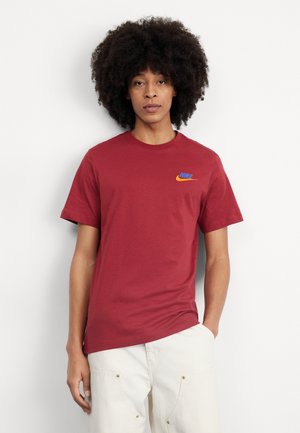 Camiseta de algodón roja de manga corta y cuello redondo, con un logo de Nike azul y naranja en la parte superior izquierda del pecho. Textura suave.