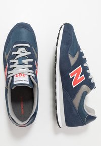 Námornické modré tenisky s vrchním materiálem z meshu a semiše, šedými akcenty, bílou mezipodešví a výrazným červeným logem 'N'. Minimalistický design a texturovaný povrch.