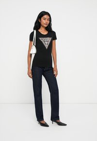 T-shirt nera in cotone con un design grafico triangolare che presenta la parola "GUESS". Abbinato a jeans dritti blu scuro e scarpe nere a punta.
