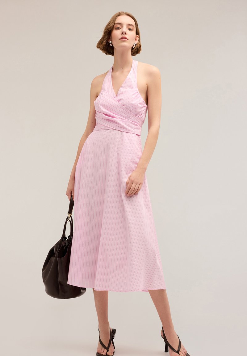 Rochie roz tip halter cu dungi verticale albe, cu un bust ajustat și o fustă midi fluidă. Modelul poartă o geantă de umăr neagră și sandale negre.