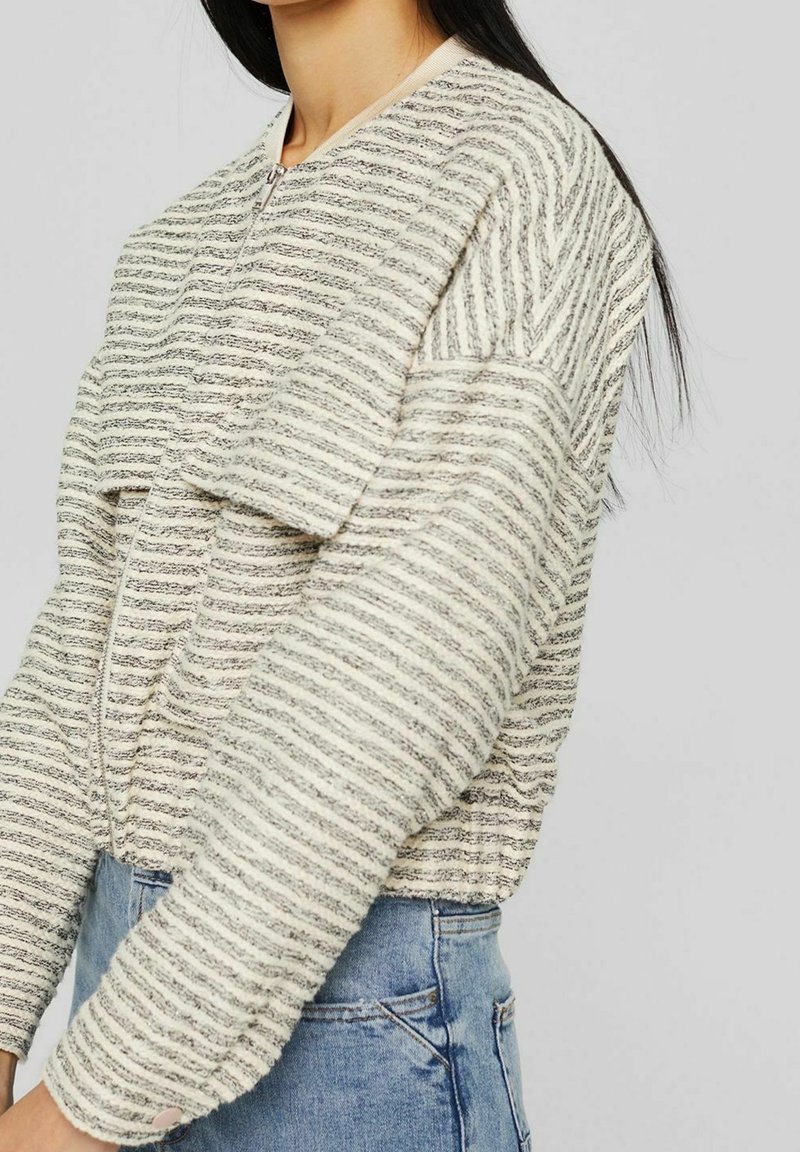 Femme portant un pull texturé rayé crème et gris à manches longues avec une fermeture éclair sur le devant et un jean en denim bleu, présenté des épaules jusqu'aux hanches.