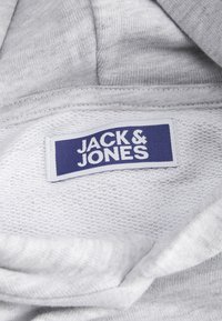 Grått textilmerke med "JACK & JONES" i vit text på en blå bakgrund. Omliggande textur är mjuk och vävd.