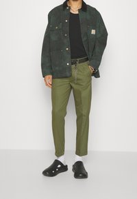 Veste verte foncée avec un col noir, deux poches avant, t-shirt noir, pantalon vert olive, chaussettes blanches et sabots noirs.