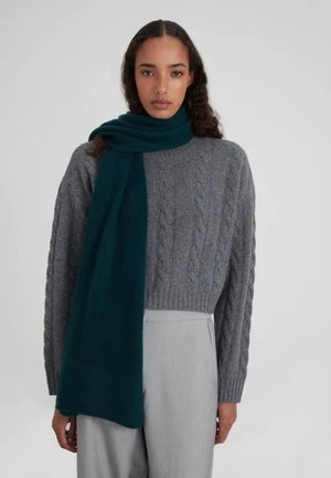 Gebreide grijze kabeltrui gecombineerd met een lange, effen teal sjaal. De sjaal hangt nonchalant over de schouders. Soepele stoftextuur.