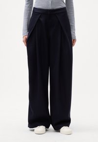 Calças largas azul-marinho com um design de frente pregueada, textura de tecido suave, cintura alta e bolsos laterais, combinadas com sapatos brancos.