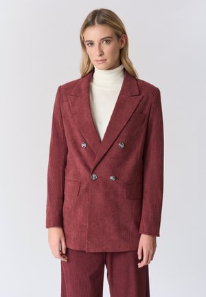 Blazer doppiopetto in velluto a coste color bordeaux con rever a punte, due tasche frontali e bottoni metallici, indossato da una persona.
