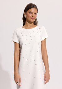 Kobieta ubrana w białą sukienkę z krótkim rękawem ozdobioną rozproszonymi perłami i cyrkoniami, do tego duże białe kolczyki i efektowny pierścionek.