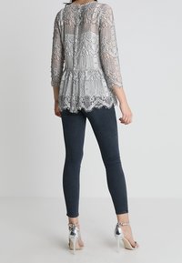 Grå spetspeplumtop med trekvartslång ärm, prydd med intrikata blommönster och en vågformad nederkant, ihop med mörka jeans och silvriga klackar.