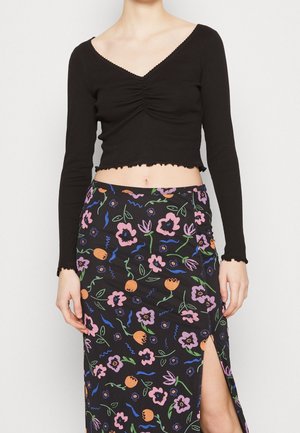 Žena v černém krátkém topu s dlouhým rukávem a vysokým pasem květinové sukně s předním rozparkem na jednobarevném pozadí.