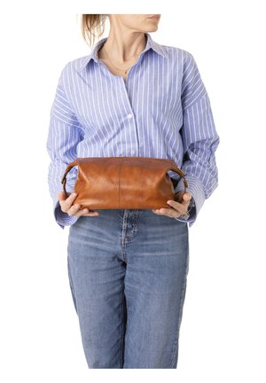 Vrouw met een blauw gestreept shirt en spijkerbroek houdt een middelgrote bruine leren tas met handvatten vast voor een witte achtergrond.