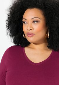 Top a V scollo bordeaux realizzato in tessuto morbido e elasticizzato. Presenta una vestibilità aderente e maniche corte, con una sottile cucitura decorativa attorno al colletto.