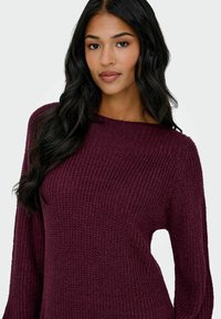 Giovane donna con lunghi capelli scuri che indossa un maglione a maglia spesso di un profondo viola su uno sfondo chiaro e semplice.