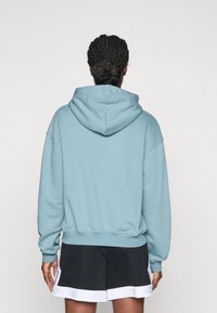 Ljusblå hoodie med dragsko i huvan, avslappnad passform och ribbade muddar; bärs tillsammans med svarta shorts med vit nederkant.