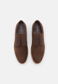 Scarpe in suede marrone con una texture liscia, presentano una punta arrotondata, dettagli cuciti e lacci grigio scuro. Soletta morbida visibile.