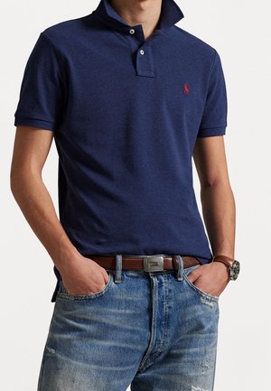 Mężczyzna w granatowej koszulce polo z czerwonym logo, niebieskich dżinsach, brązowym pasku i zegarkiem na nadgarstku, z rękami w kieszeniach, na jednolitym tle.