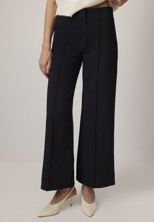 Nő magasderekú, fekete, bő szárú nadrágban, fehér, hegyes orrú, pántos magassarkú cipőkkel, semleges háttér előtt.