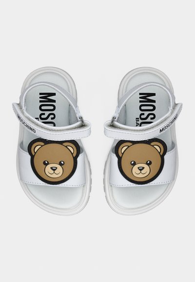 Λευκά σανδάλια παιδικά με πρόσωπα αρκουδάκια στα μπροστινά λουράκια και ρυθμιζόμενα λουράκια αστραγάλου με τη μάρκα "Moschino".