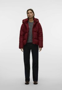 Rød polstret jakke med hætte, front lynlås, sidelommer, båret over en grå sweater og mørkeblå jeans, stående mod en ensfarvet baggrund.
