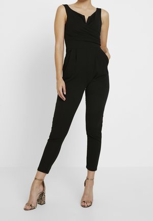 Kvinde iført en sort ærmeløs jumpsuit med V-udskæring og ankellange bukser, kombineret med højhælede sandaler med slangeprint.