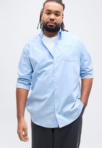 Άνδρας με γενειάδα και κοτσιδάκια, που φορά ένα γαλάζιο πουκάμισο πάνω από ένα λευκό μπλουζάκι, στέκεται με το ένα χέρι στην τσέπη και ουδέτερη έκφραση.