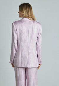 Completo rosa chiaro con una finitura testurizzata lucida, dotato di un'unica fessura posteriore, rever a punta e dettagli con bottoni sulle maniche e sui pantaloni.