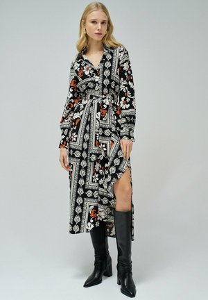 Largo vestido negro con patrones florales y geométricos en blanco y naranja, con un diseño envolvente y una abertura lateral. La modelo lleva botas negras hasta la rodilla.