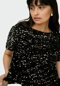 Haut noir à sequins avec un col rond, des manches courtes et une coupe ample, orné de sequins métalliques reflétant différentes nuances de lumière.