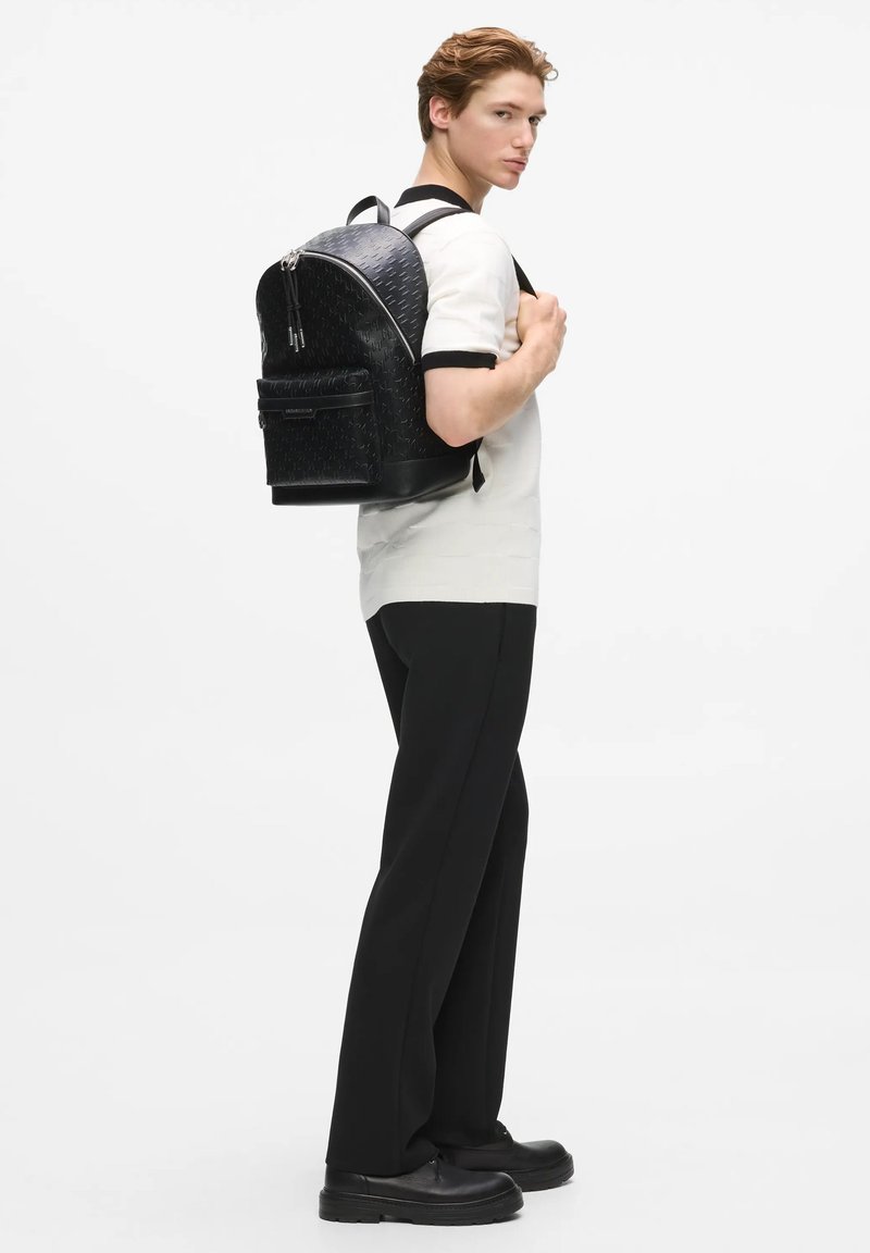 Jeune homme portant une chemise blanche à manches courtes et un pantalon noir, portant un sac à dos en cuir noir, posant de profil sur un fond blanc.