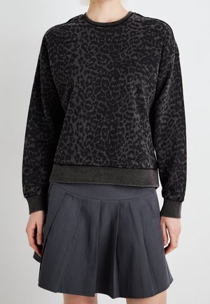 Person trägt einen langärmeligen schwarzen und dunkelgrauen Leopardenmuster-Sweatshirt und einen plissierten dunkelgrauen Rock, steht vor einem schlichten Hintergrund.