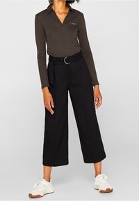 Polo a maniche lunghe di colore marrone scuro con colletto e piccolo logo, abbinato a pantaloni neri a gamba larga a vita alta e sneakers bianche.