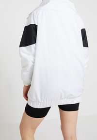 Biała kurtka z czarnymi panelami na ramionach, luźnym krojem i elastycznym pasem. Gładki materiał z kołnierzem i opuszczanymi ramionami.