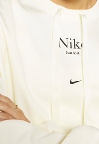Cream-colored hoodie made of soft fabric featuring a black "Nike" logo and "Just do it" text, with drawstrings and a relaxed fit.