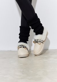 Pantofole beige trapuntate con bordino in pelliccia, cinturino a motivo bianco e nero, dettagli in metallo dorato, abbinate a scaldamuscoli neri a coste.