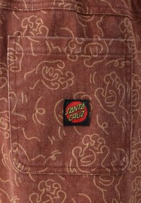 Brown fabric featuring a repeating yellow line drawing pattern. A black patch with "SANTA CRUZ" in red and green is visible.