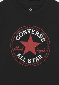 Melna kokvilnas t-krekls ar lielu sarkanu zvaigzni un baltu tekstu, kas lasāms "CONVERSE All Star" un "Chuck Taylor" apļveida dizainā.