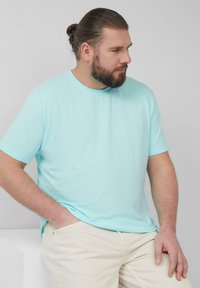 Man met een baard en teruggebonden haar draagt een lichtblauw t-shirt en beige broek, zit met één hand in zijn zak tegen een effen achtergrond.
