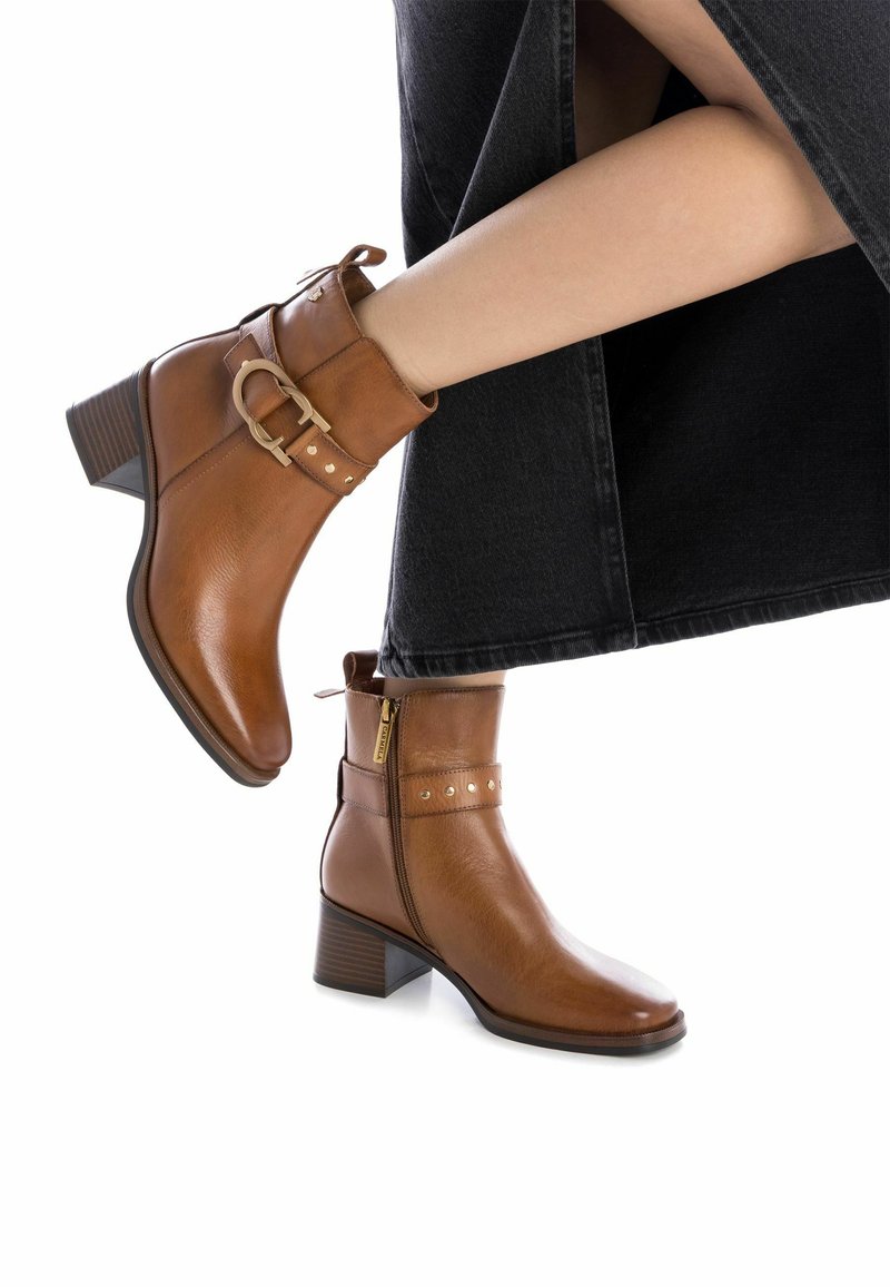 Las botas de cuero marrón hasta el tobillo presentan una punta redonda, un zipper lateral, tachuelas decorativas y un tacón grueso con un acabado texturizado.