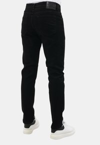 Persoon in slim-fit zwarte jeans met achterzakken en witte sneakers met zwarte hielaccenten, staand tegen een effen achtergrond.