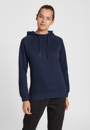 Kobieta w granatowej bluzie z kapturem i ściągaczami oraz czarnych spodniach, stojąca na jednolitym jasnoszarym tle.