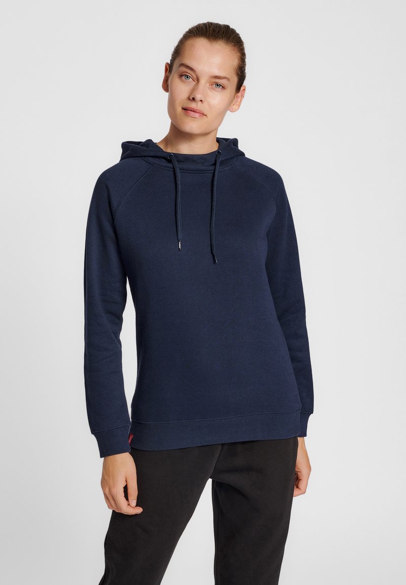 Kobieta w granatowej bluzie z kapturem i ściągaczami oraz czarnych spodniach, stojąca na jednolitym jasnoszarym tle.