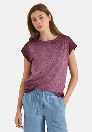 Jeune femme aux cheveux bruns portant un haut violet à manches courtes et un pantalon bleu clair à cordon, debout devant un fond uni.