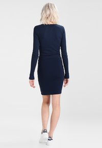 Námořnické modré šaty s dlouhým rukávem a přiléhavým střihem, hladká textura a kulatý výstřih, doplněné bílými teniskami.