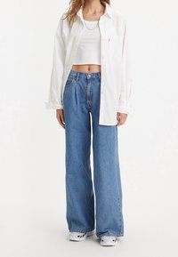 Vit långärmad skjorta över en vit kortärmad topp, i kombination med löst sittande blå denimjeans och vita sneakers. Skjortan har en liten ficka.