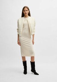 Quilted cream bomber jacket over a form-fitting, knee-length beige dress, paired with tall black heeled boots. Smooth textures and subtle details.